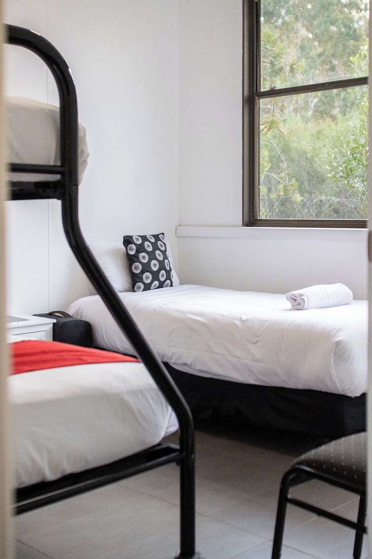 A dormitory room with two beds, a window, and a chair.