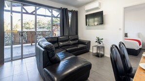 A modern living room with a black leather sofa, a flat-screen TV mounted on the wall, and a dining area with a wooden table and chairs.