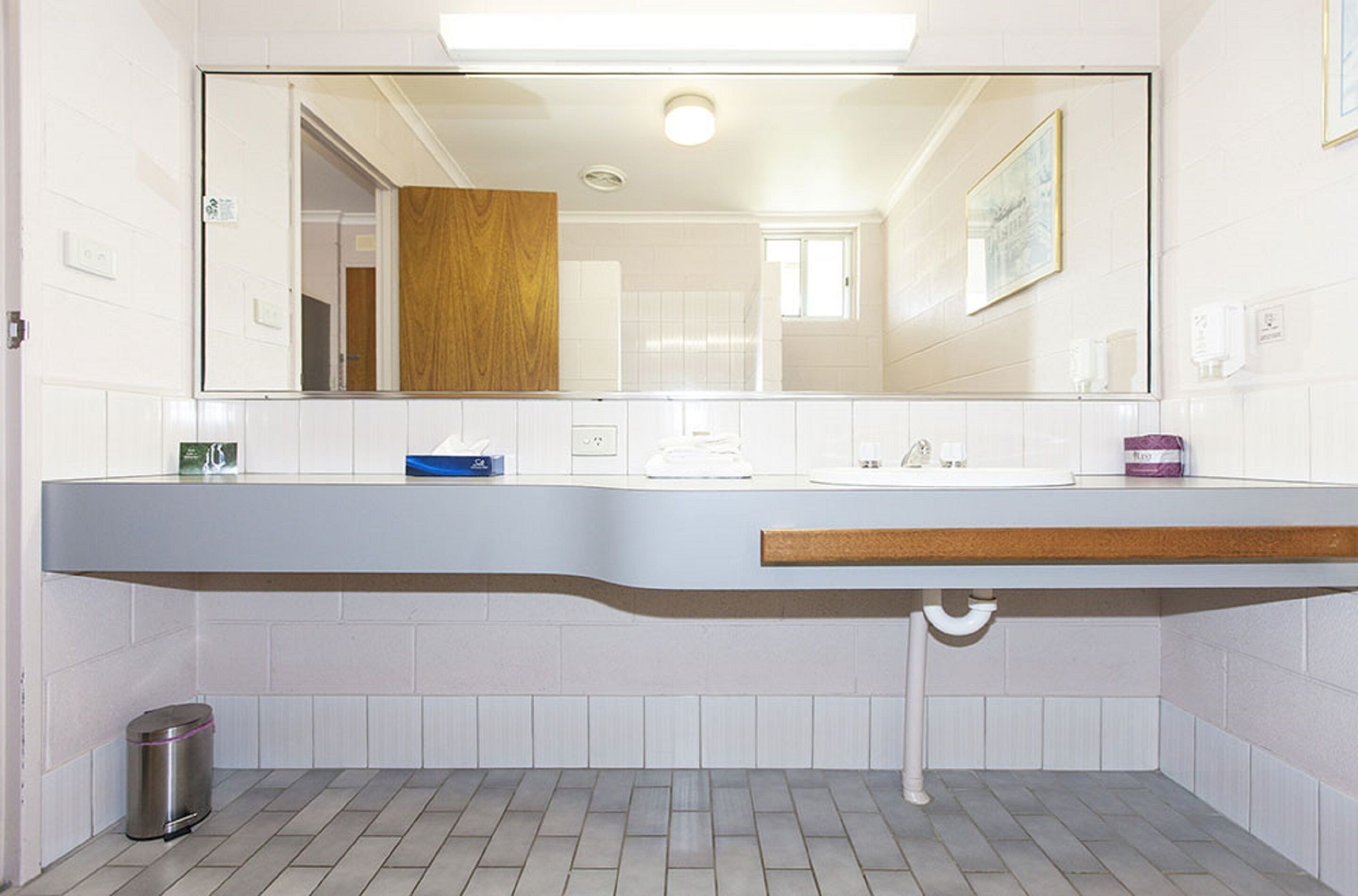 Un baño con pared de azulejos blancos, encimera de madera, lavamanos, espejo, cubo de basura y ventana.
