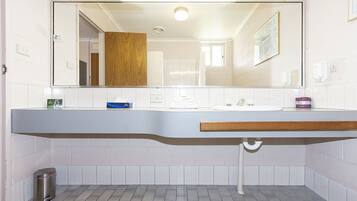 Un baño con pared de azulejos blancos, encimera de madera, lavamanos, espejo, cubo de basura y ventana.