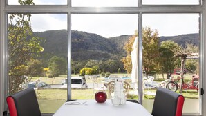A dining area with a table set for two, a red decorative item, and a menu stand. Outside, there is a view of a park with trees, a bicycle, and a car.