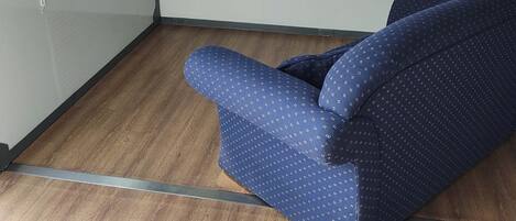 A compact room with a blue patterned sofa, a flat-screen TV mounted on the wall, and a window with a view of greenery.