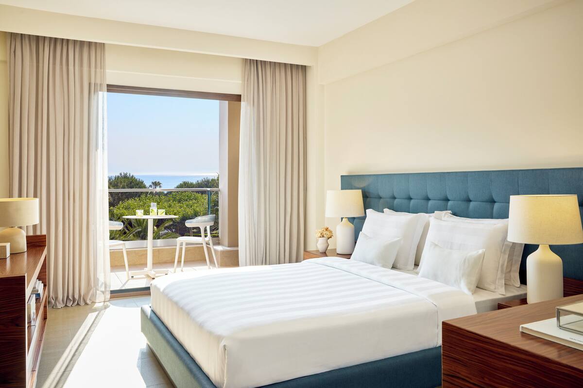 A hotel room with a large bed, two bedside tables with lamps, a view of the outdoors, and a balcony with a table and chairs.