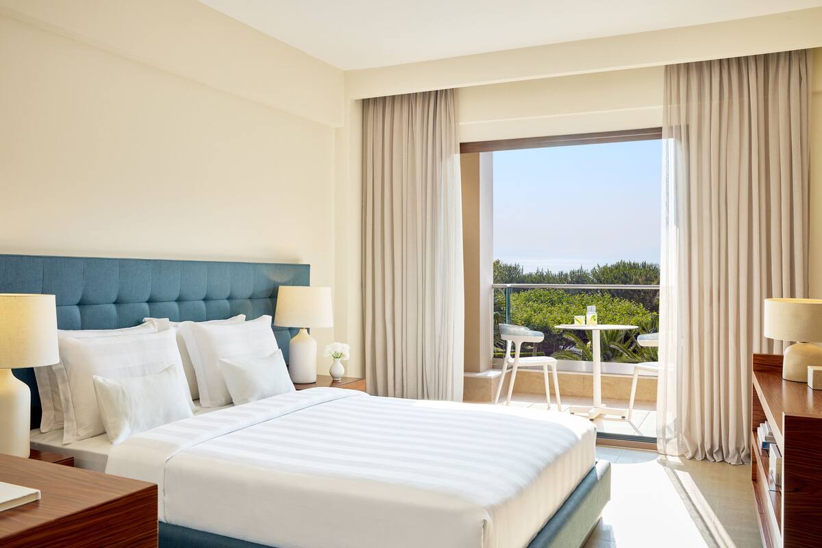 A hotel room with a large bed, a blue headboard, two bedside lamps, a small table with a chair, and a balcony with a view of greenery.
