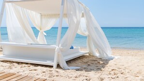 A white canopy bed on a sandy beach with clear blue water in the background. - Pomegranate Wellness Spa Hotel (Nea Propontida)