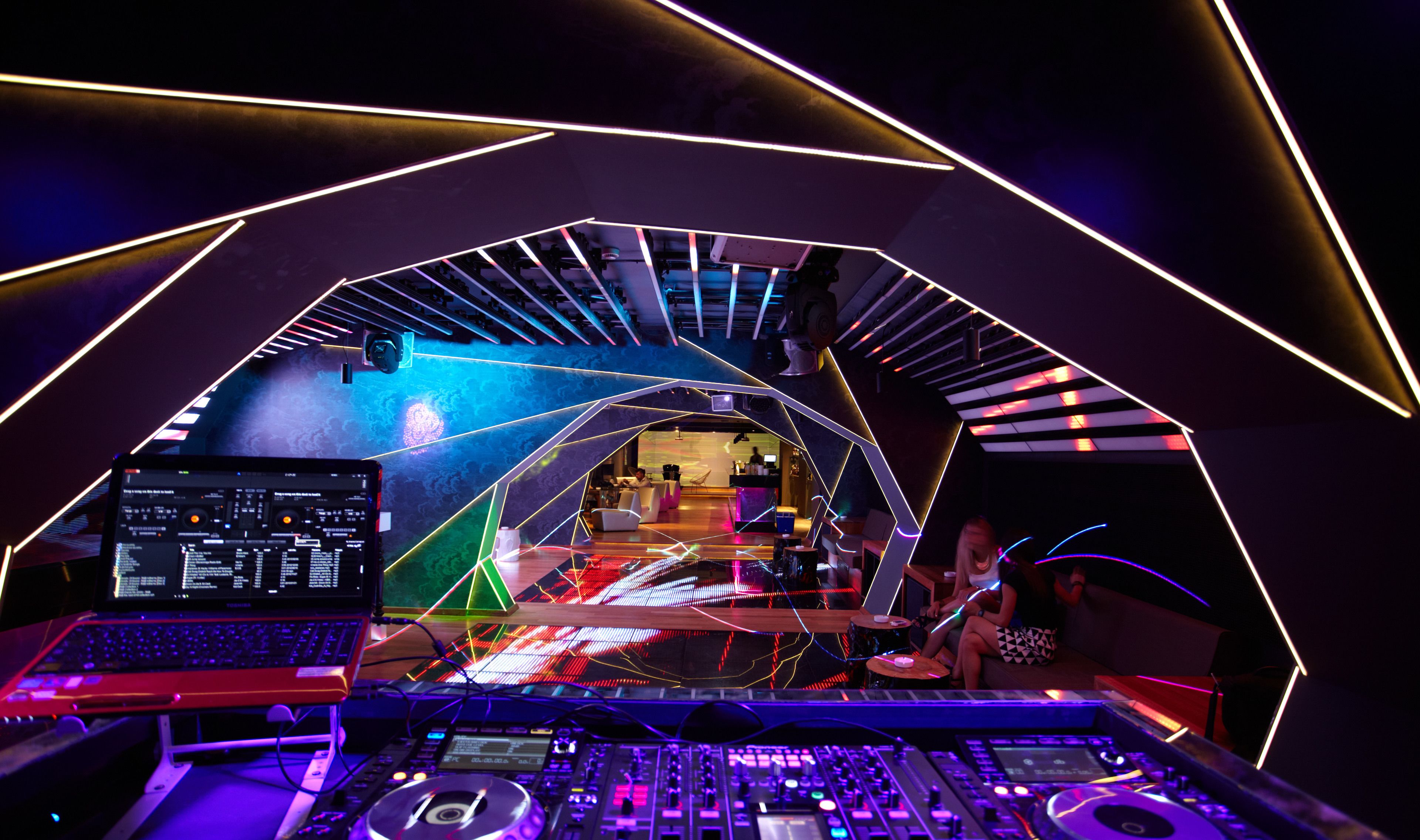 A modern DJ booth with a laptop, mixer, and turntables, surrounded by neon lights and geometric patterns.