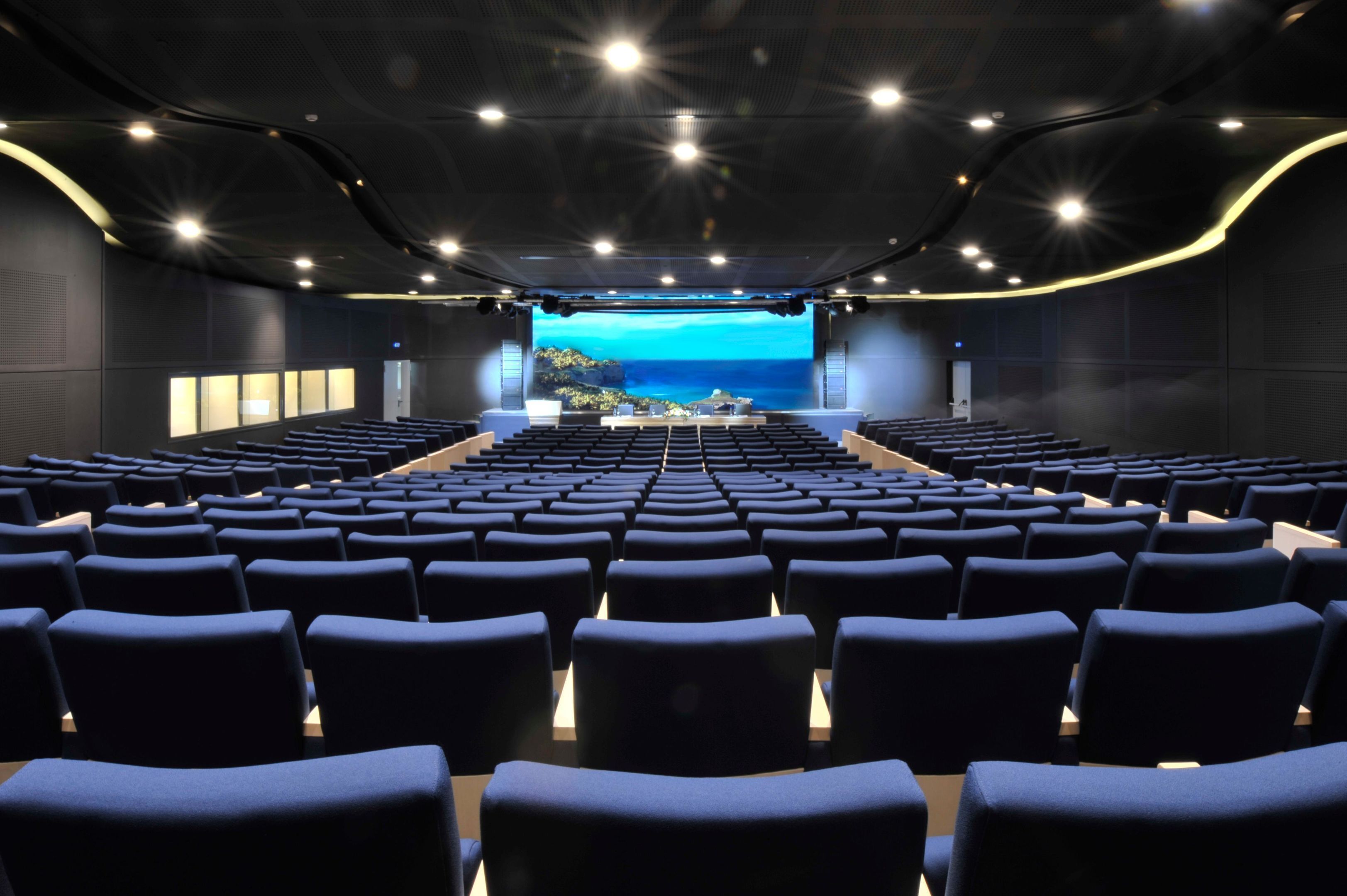 A theater with rows of blue seats, a stage with a scenic backdrop, and a modern ceiling design.