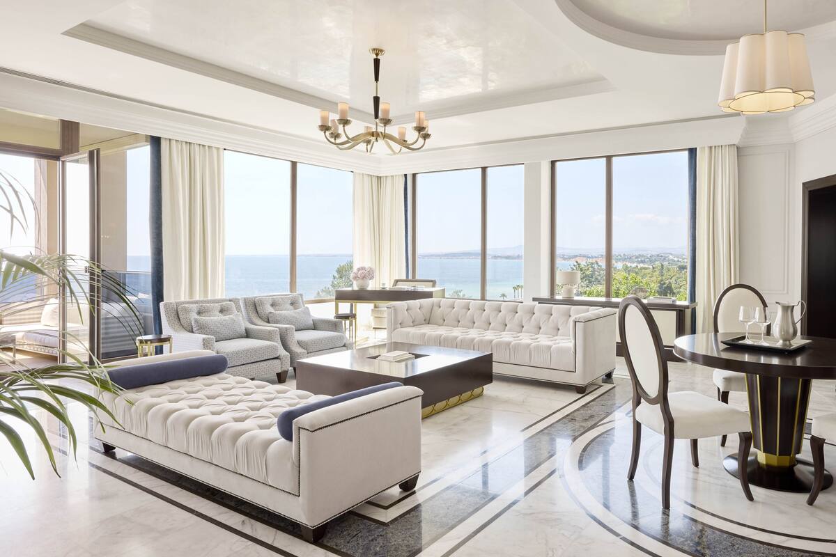 A spacious living room with a large sofa, a coffee table, and a dining table with chairs. There is a chandelier and a view of the sea through large windows.