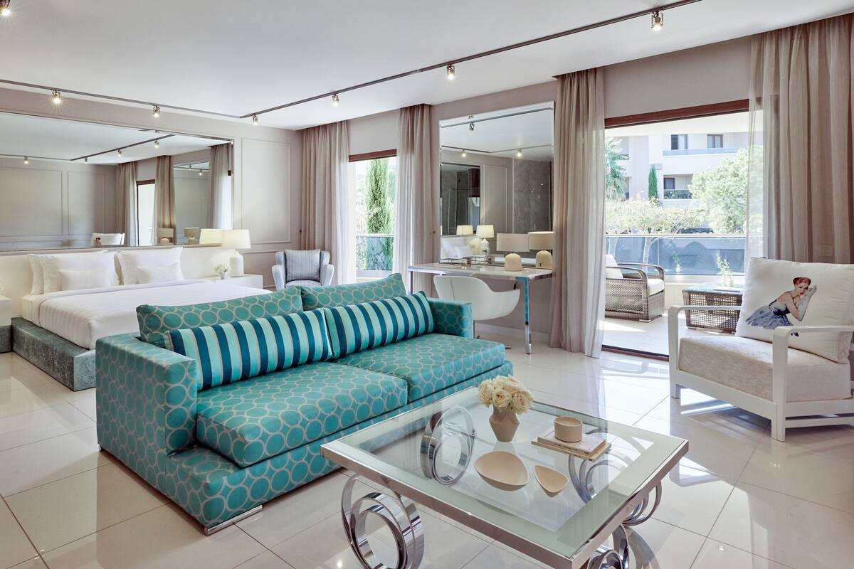 A modern hotel room with a large bed, a sofa, a glass coffee table, and a balcony with a view.