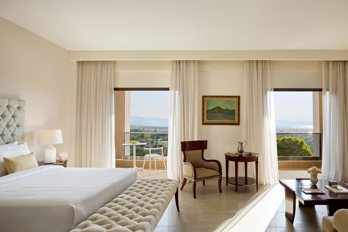 A hotel room with a large bed, a chair, a small table, and a view of the landscape through the windows.