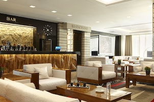 Un moderno vestíbulo de hotel con zona de bar y restaurante, con sofás, mesas y un reloj.