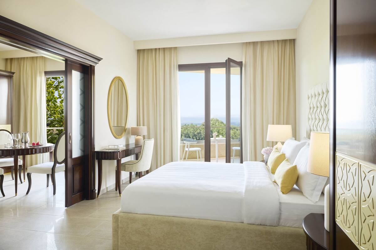A hotel room with a large bed, a dining area with a table and chairs, a view of the outdoors, and a balcony with a table and chairs.