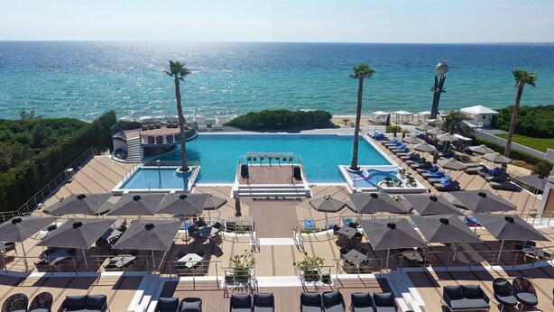 A pool area with lounge chairs and umbrellas, surrounded by palm trees and overlooking the sea. - Pomegranate Wellness Spa Hotel (Nea Propontida)