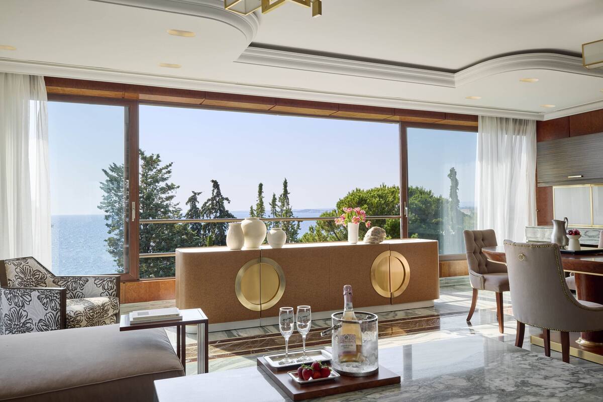A luxurious room with a large window, a marble floor, a wooden sideboard, and a dining area with a table and chairs.
