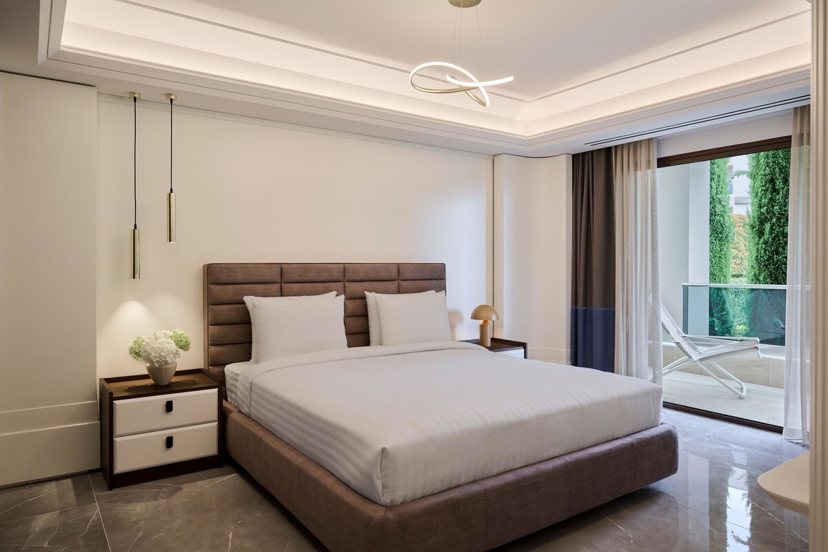 A modern bedroom with a large bed, a nightstand with a lamp, and a view of the outdoors through a sliding glass door.