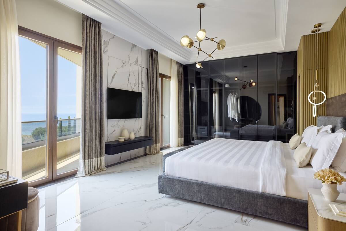 A modern bedroom with a large bed, a TV, and a view of the sea.