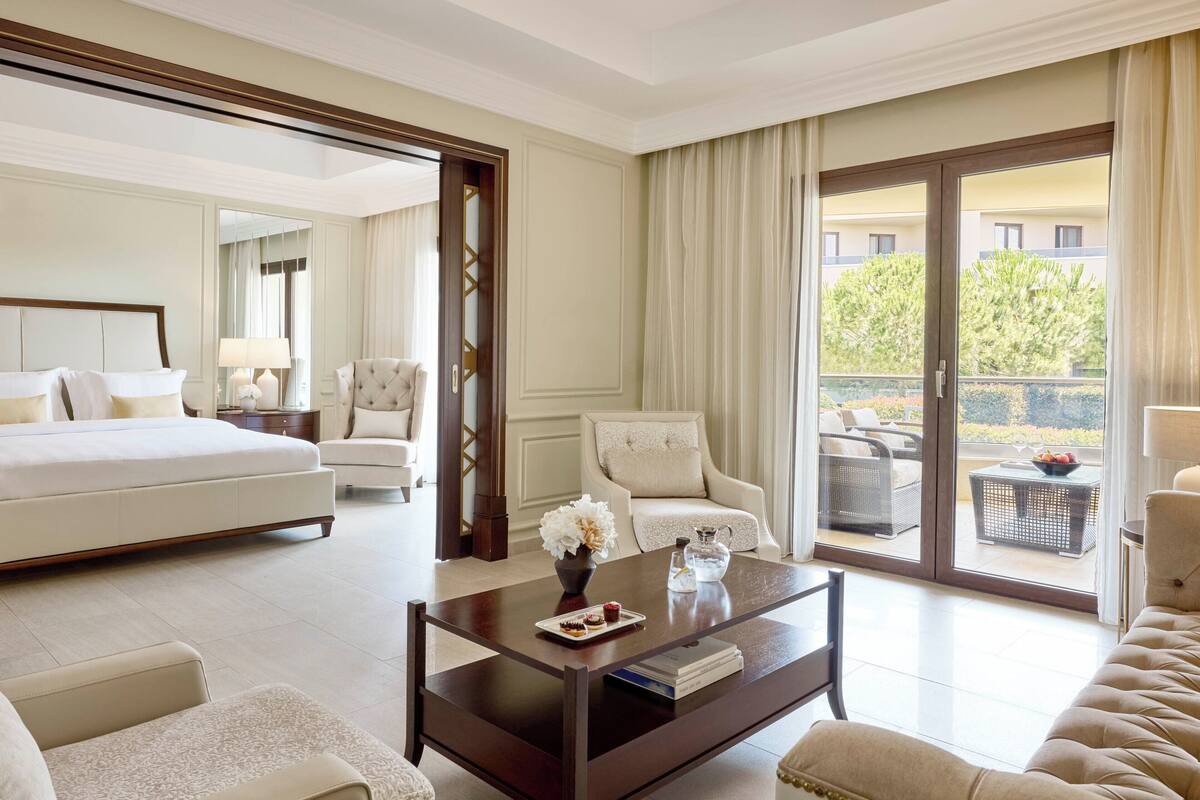 A hotel room with a large bed, a sitting area with a sofa and armchairs, a coffee table with a vase and book, and a view through the glass door.