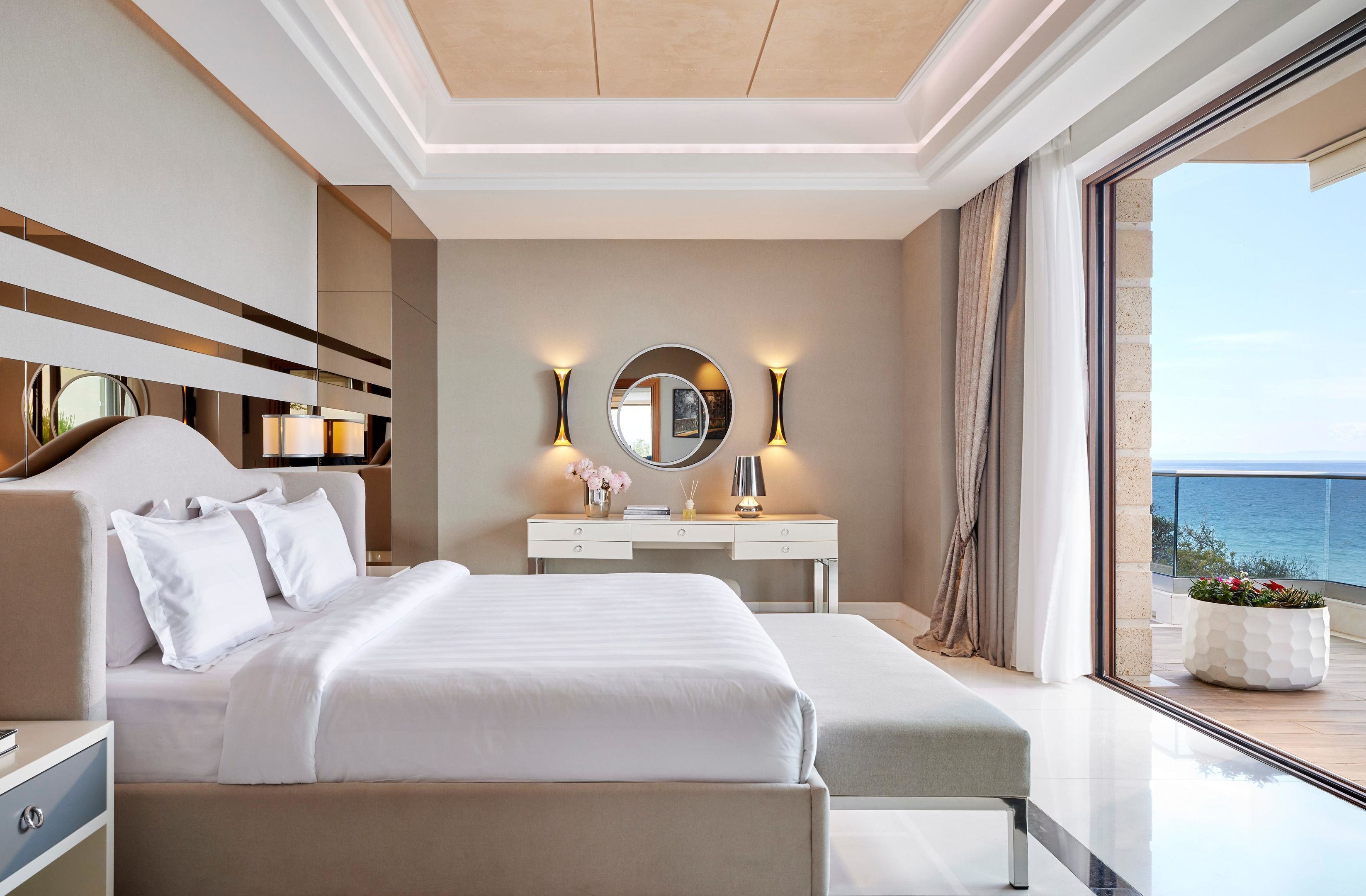 Una habitación de hotel moderna con una cama grande, una mesita de noche, un tocador con espejo y vistas al mar.
