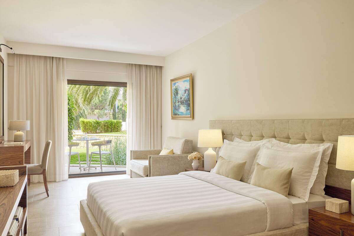A hotel room with a large bed, two bedside tables with lamps, a desk with a chair, and a view of a garden through a glass door.
