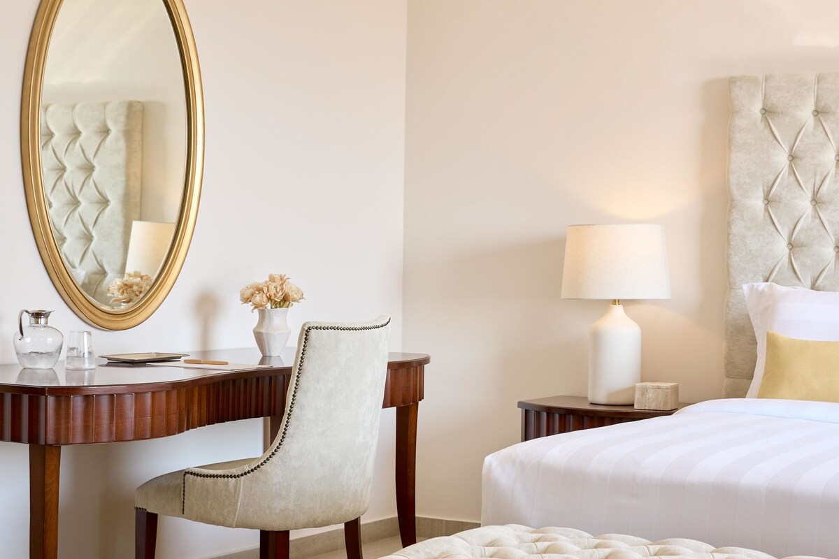 A hotel room with a bed, a desk with a chair, a lamp, a mirror, and a vase with flowers.