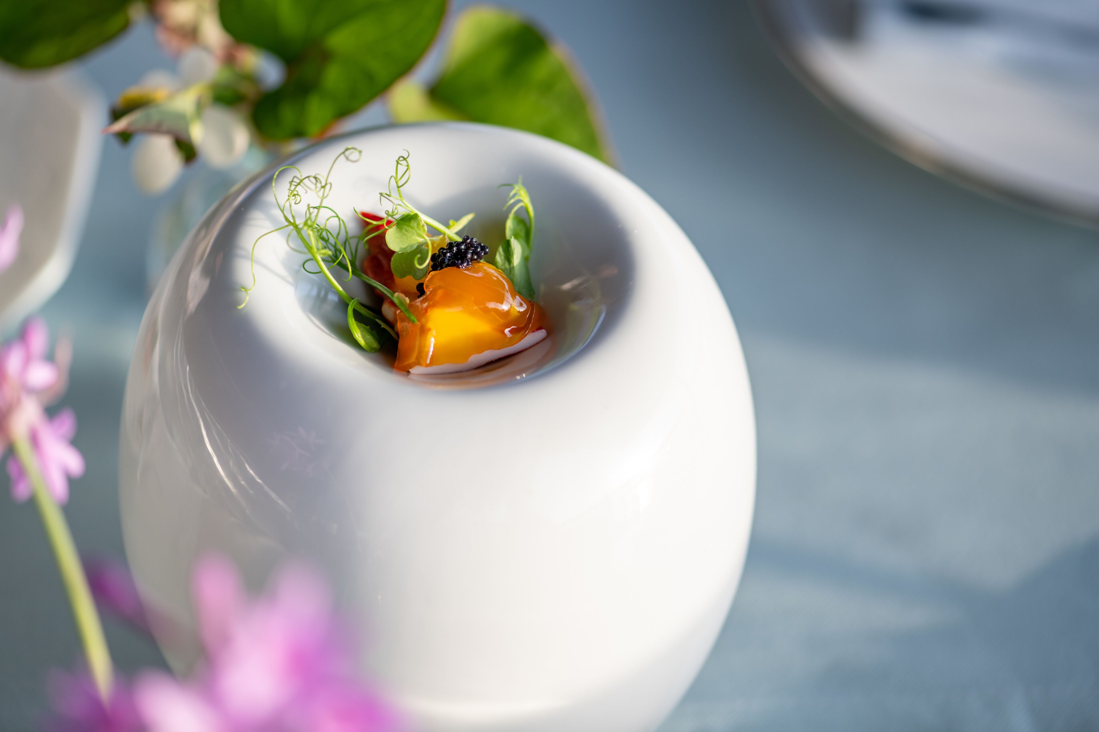 Un bol blanc contenant un dessert, décoré de quelques feuilles vertes et d’une baie, sur une table. L’arrière-plan est flou.