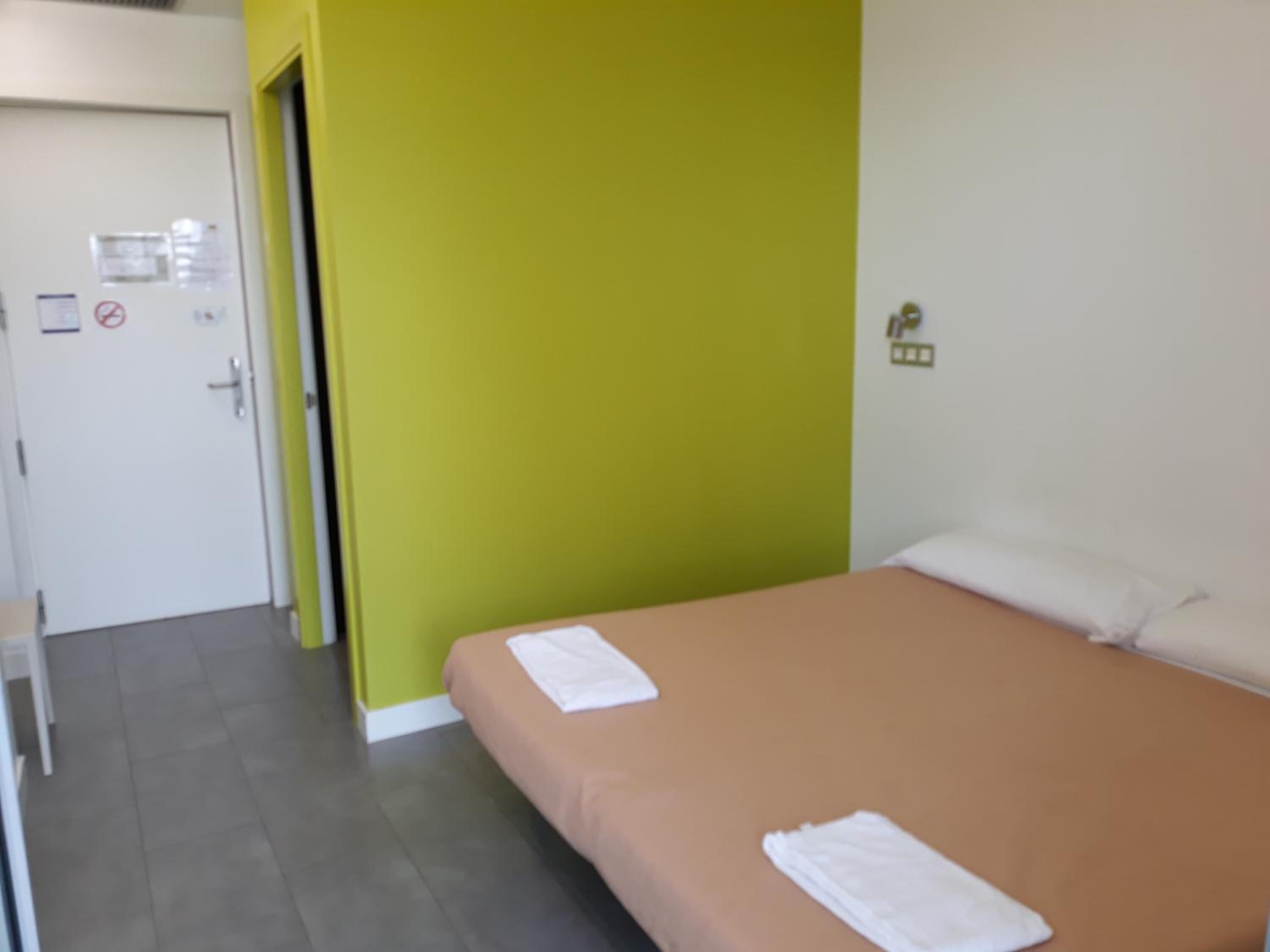 Un lit simple avec un matelas blanc et deux oreillers, une porte sur laquelle est apposé un panneau « interdiction de fumer », et un mur de couleur vert clair.