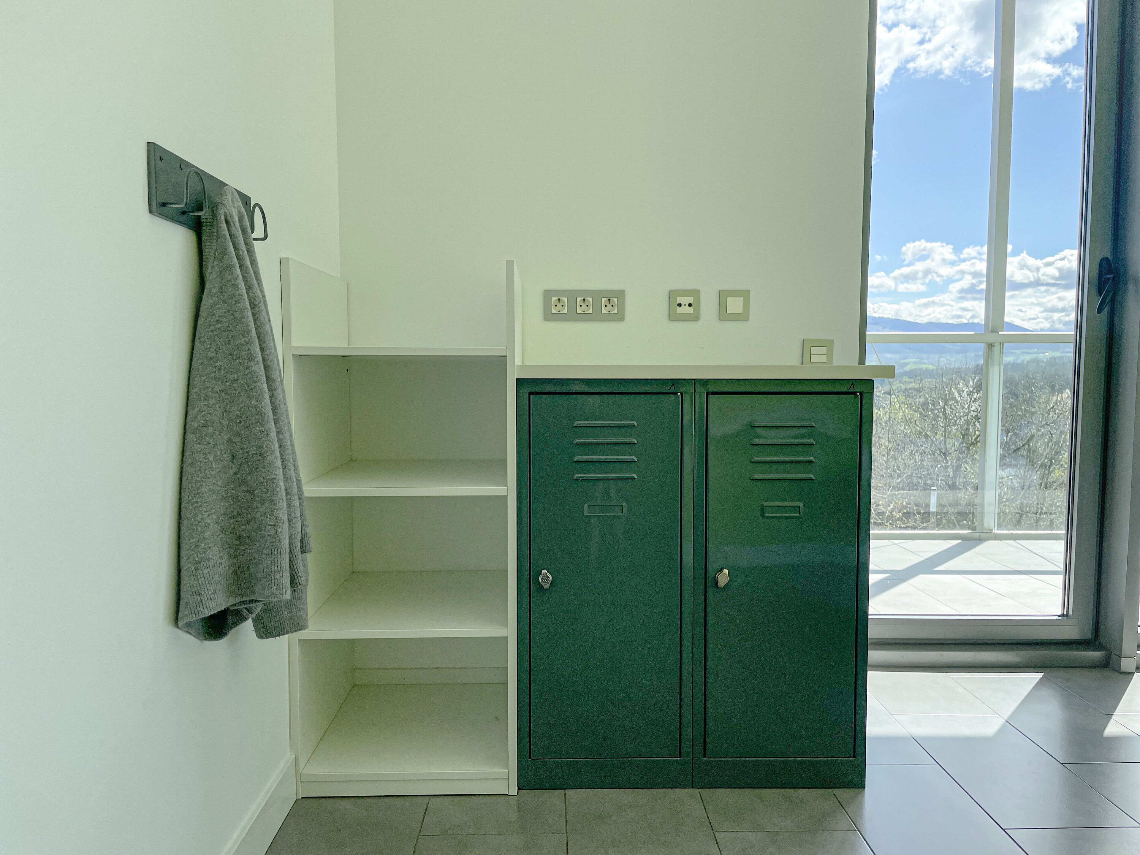 Una habitación con un casillero verde, una toalla colgada de un perchero y un ventanal con vistas a árboles y montañas.