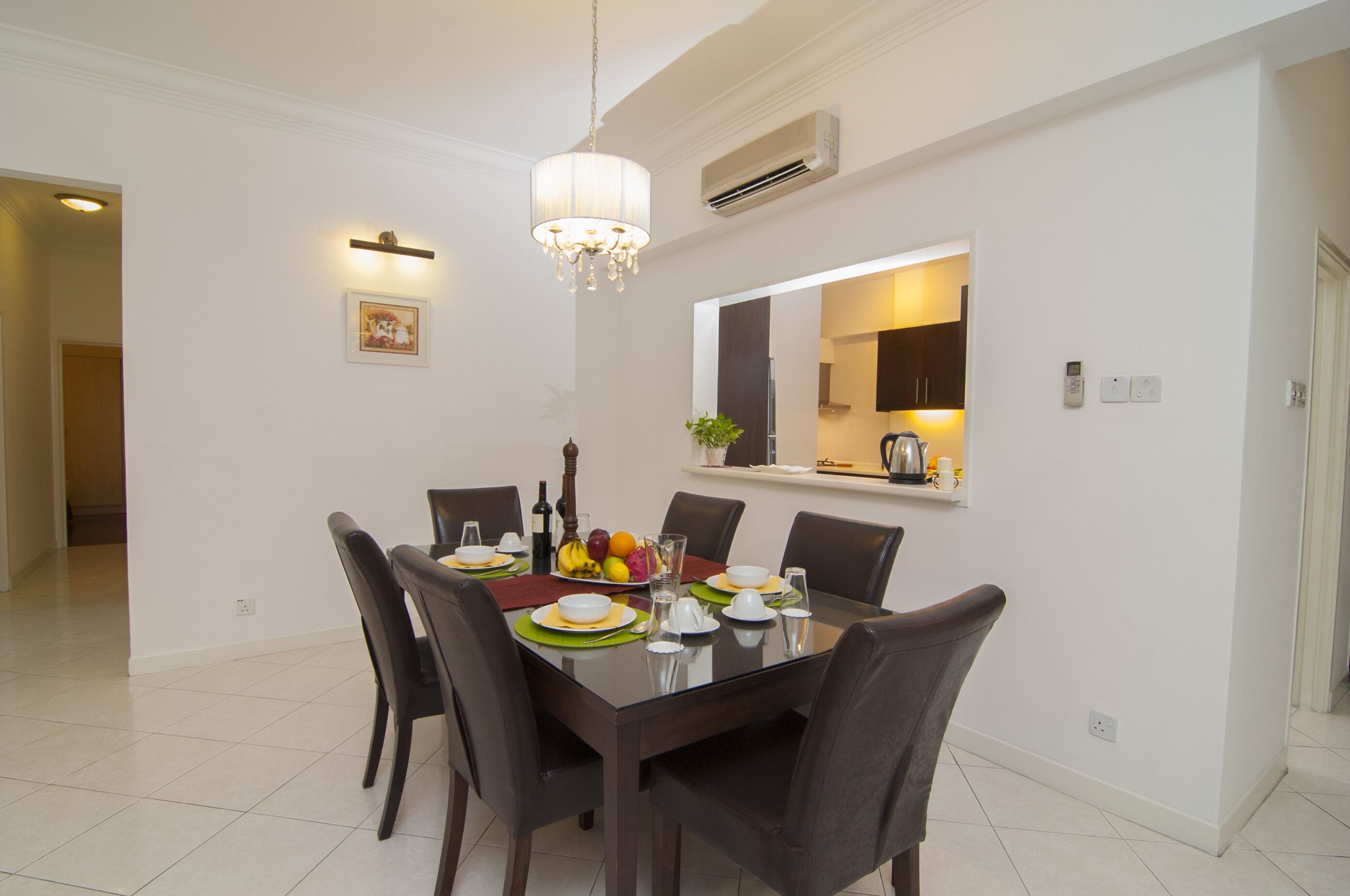 Un comedor con una mesa rectangular dispuesta para seis personas, rodeada de sillas oscuras. Un candelabro cuelga sobre la mesa y un equipo de aire acondicionado está montado en la pared.