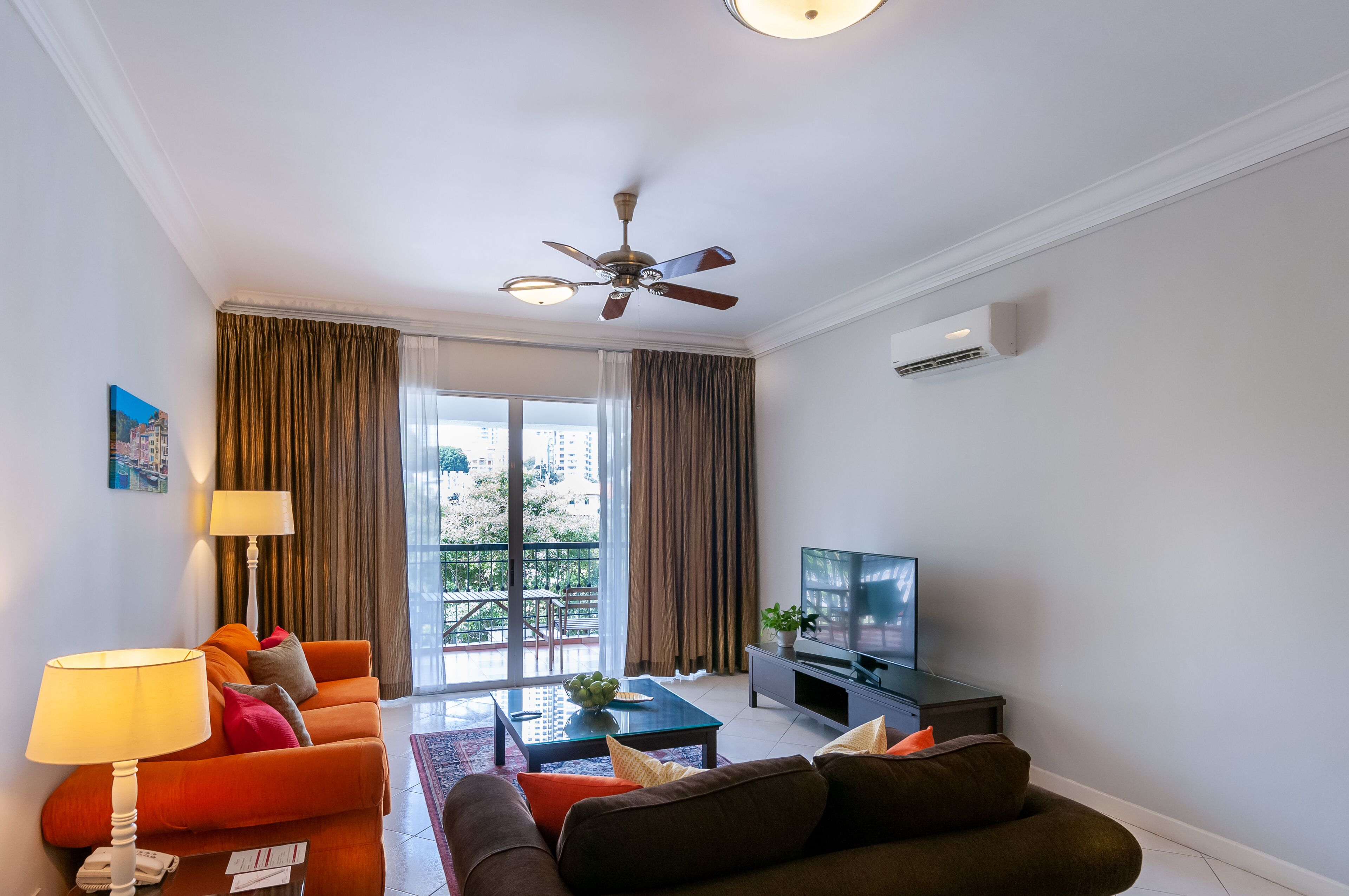 A modern living room with a sofa, armchair, and a television. There is a ceiling fan, a window with curtains, and a view of buildings outside.
