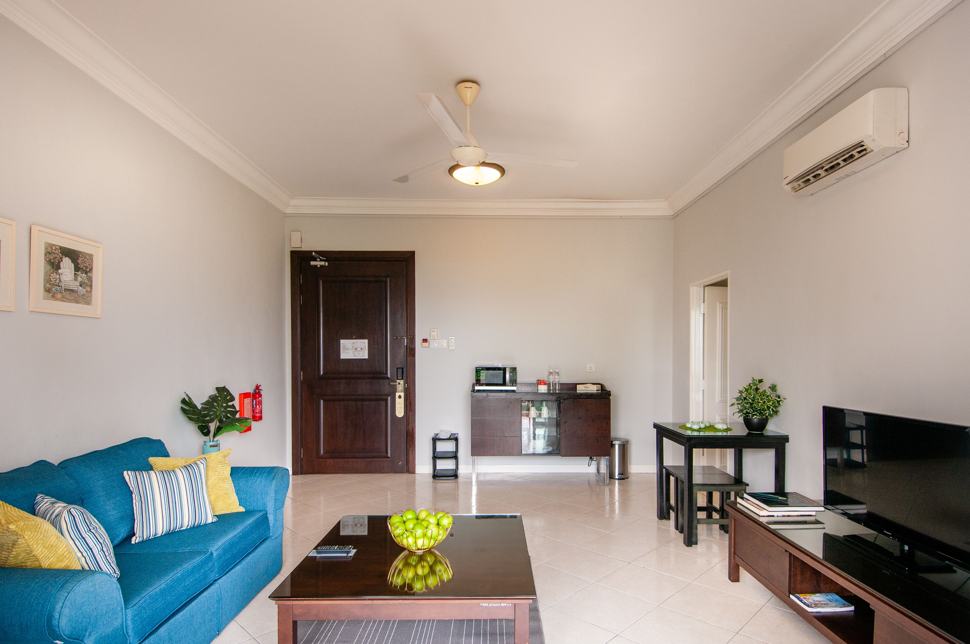 A living room with a blue sofa, a coffee table with a fruit bowl, a TV, and a kitchen area in the background.