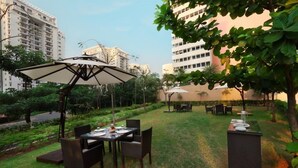 Un área de comedor al aire libre con mesas y sillas, rodeada de follaje y edificios al fondo.