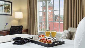 Una habitación de hotel con una cama, una bandeja con gofres y fruta, dos vasos de jugo de naranja, y vistas a un edificio rojo con una torre del reloj.