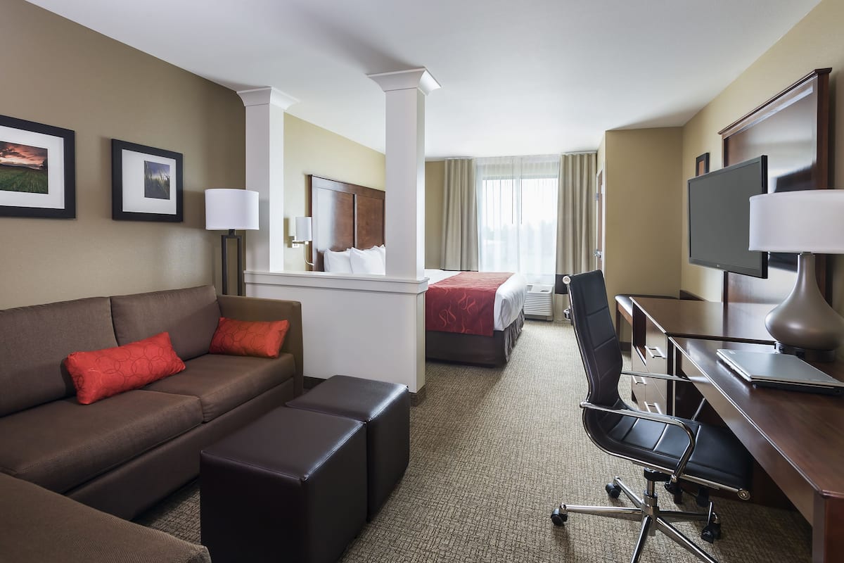 Une chambre d’hôtel avec un canapé marron, des coussins rouges, un bureau avec un ordinateur, une chaise, un lit avec une couverture rouge et deux tableaux encadrés au mur.