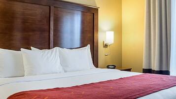 A hotel room with a large bed, a wooden headboard, a red throw blanket, a bedside table with a lamp, and a window with curtains.
