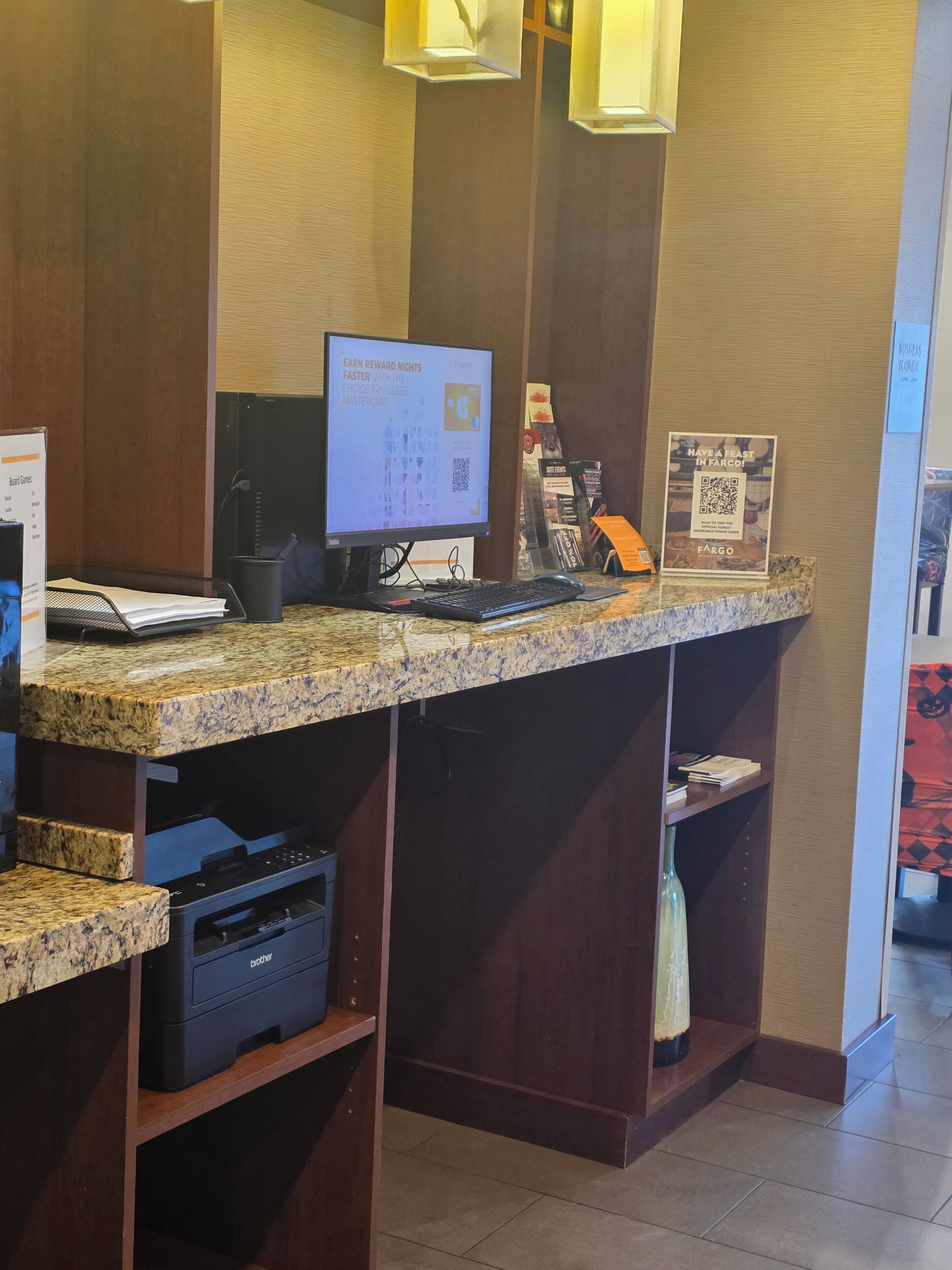A hotel reception desk with a computer monitor displaying a rewards program, a printer, and a sign promoting a local event.