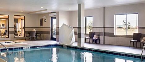 An indoor swimming pool with clear water, a tiled floor, and a few chairs and tables near the pool.