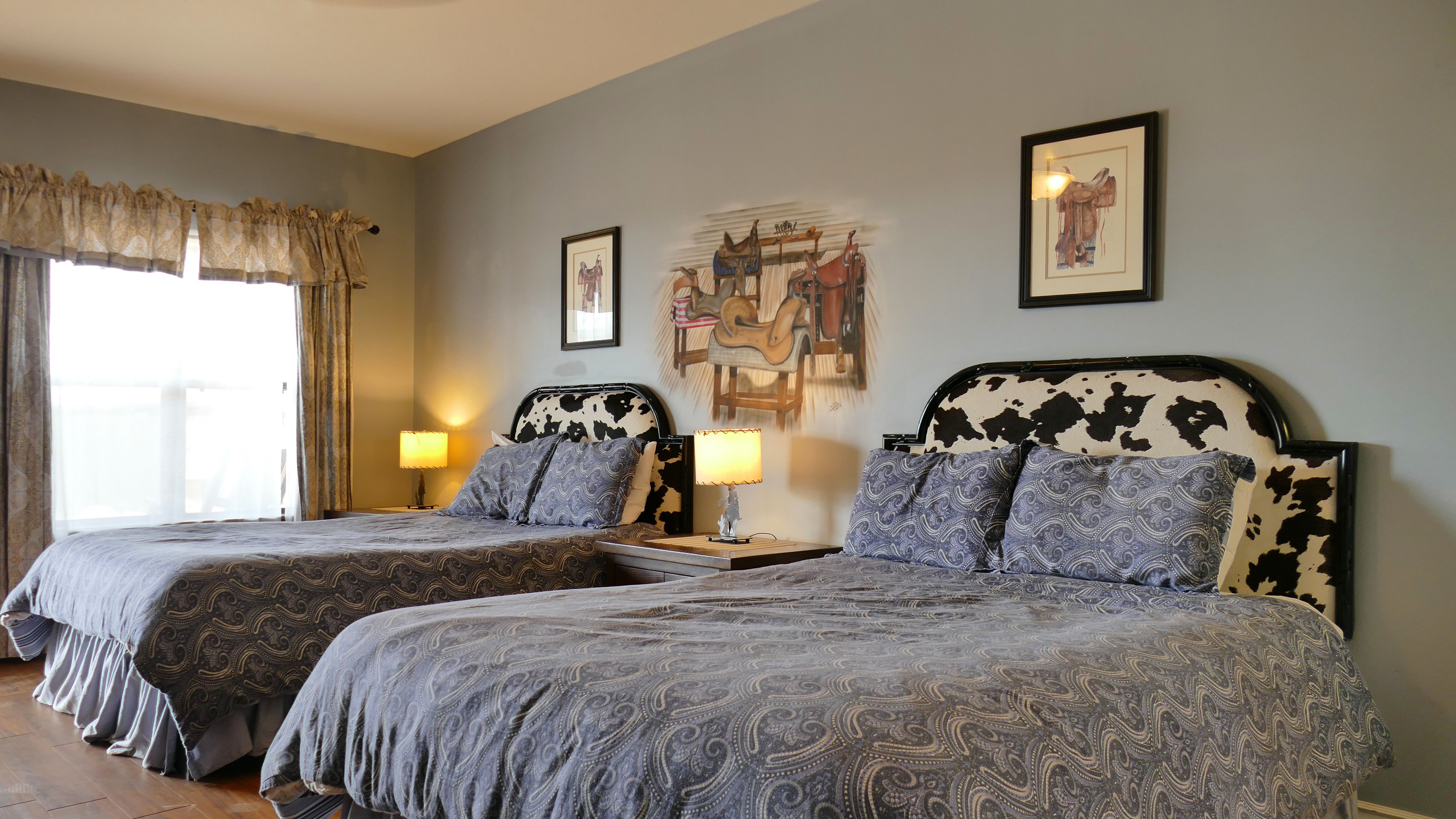 A hotel room with two beds, each with a headboard featuring a cow print design. There are two bedside tables with lamps, and the walls are adorned with framed artwork.