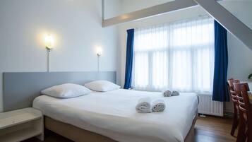 A neatly made bed with white linens and two pillows, a bedside table, a chair, and a window with blue curtains.