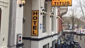 L’entrée d’un hôtel, avec une enseigne indiquant « HÔTEL TITUS » et une autre enseigne, plus petite, au-dessus de la porte, portant le mot « HÔTEL ». Des marches mènent à l’entrée, et des vélos sont garés à l’extérieur.
