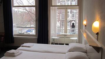 Ein Hotelzimmer mit zwei Betten, einem Tisch und einem Stuhl. Es gibt Fenster mit Vorhängen, eine Heizung und eine Wandleuchte.