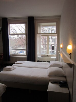 A hotel room with two beds, a table, and a chair. There are windows with curtains, a radiator, and a wall-mounted light fixture. - Hotel Titus (Amsterdam)