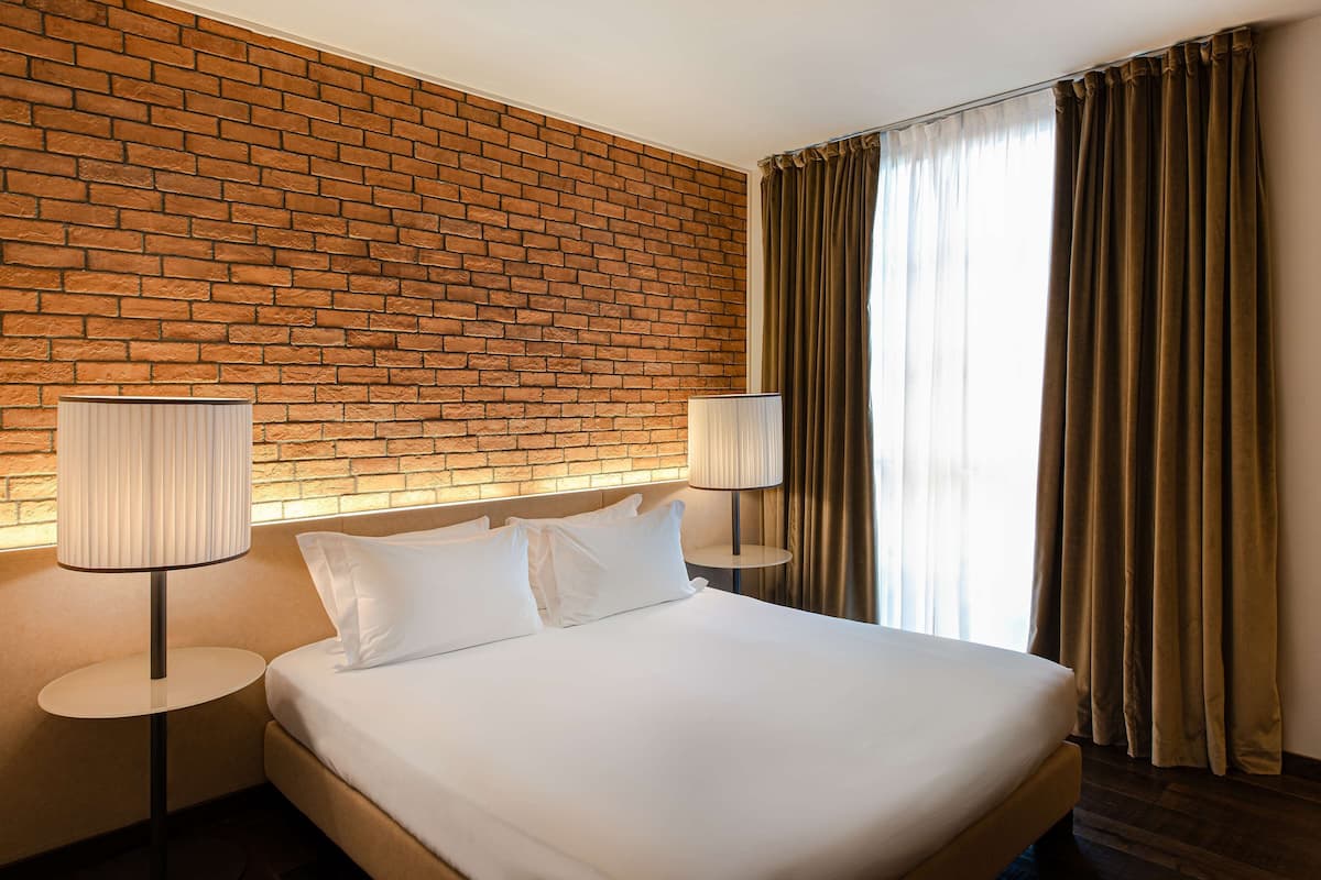 Une chambre d’hôtel avec un mur décoré d’un motif en briques, un grand lit avec une literie blanche, deux lampes de chevet et une fenêtre avec des rideaux.
