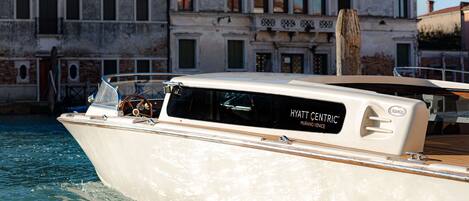 Un yate blanco con la inscripción "HYATT CENTRIC MURANO VENICE" en el costado, amarrado en un canal con edificios históricos al fondo.