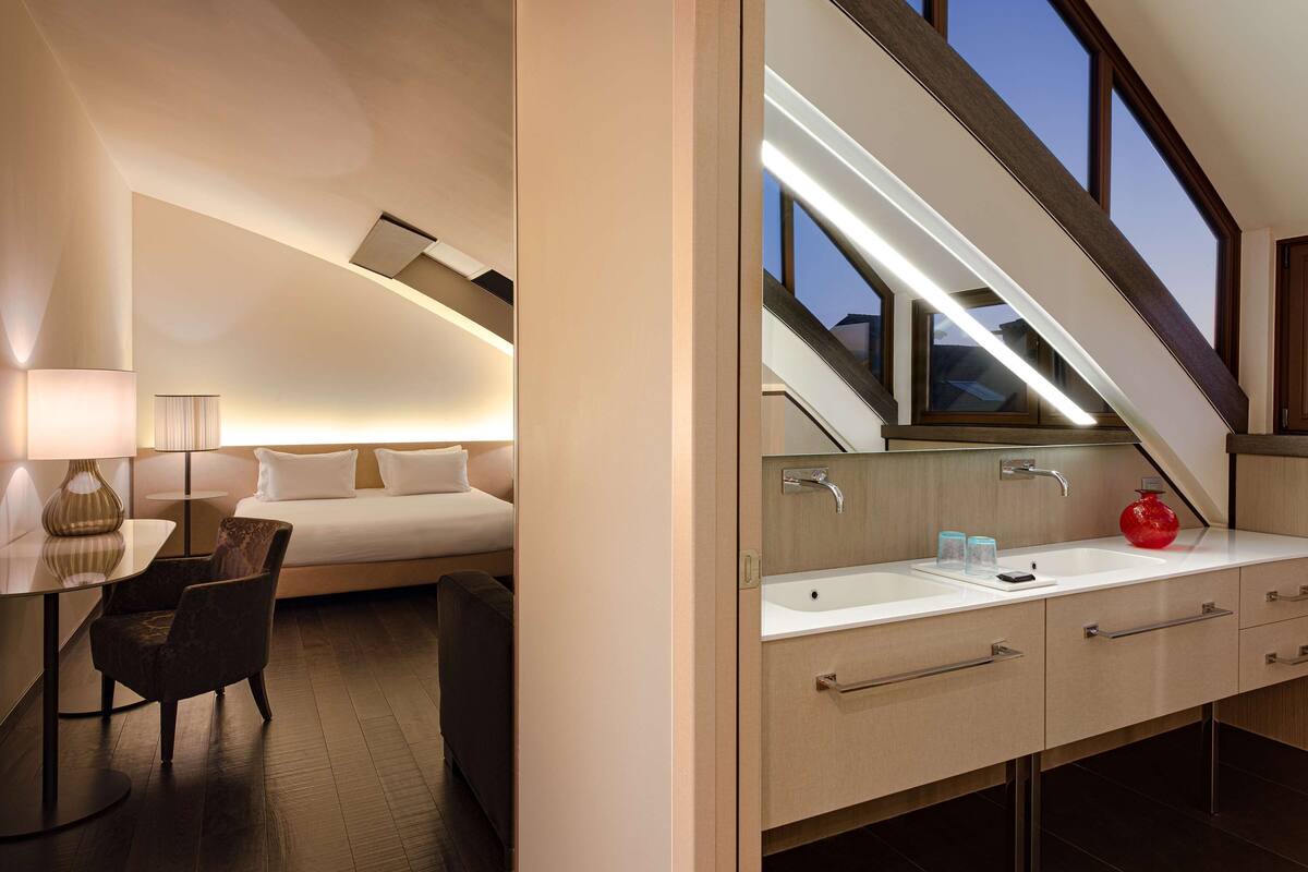 Une chambre d’hôtel moderne, équipée d’un lit, d’un bureau avec une lampe, d’une salle de bain avec un lavabo et un miroir, et offrant une vue sur le ciel par une fenêtre.