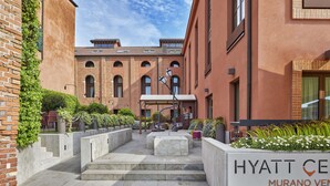 Un patio con un sendero de piedra que lleva a una zona de estar, un letrero del hotel Hyatt Centro Murano Venezia, y edificios de ladrillo al fondo.
