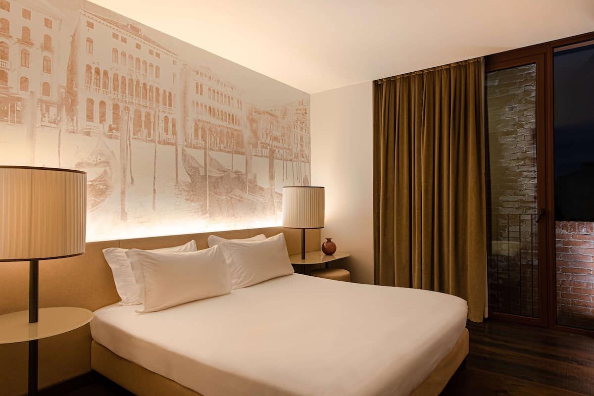 Une chambre d’hôtel avec un grand lit, deux tables de chevet avec des lampes, une grande fresque murale et une porte-fenêtre coulissante donnant sur un balcon.