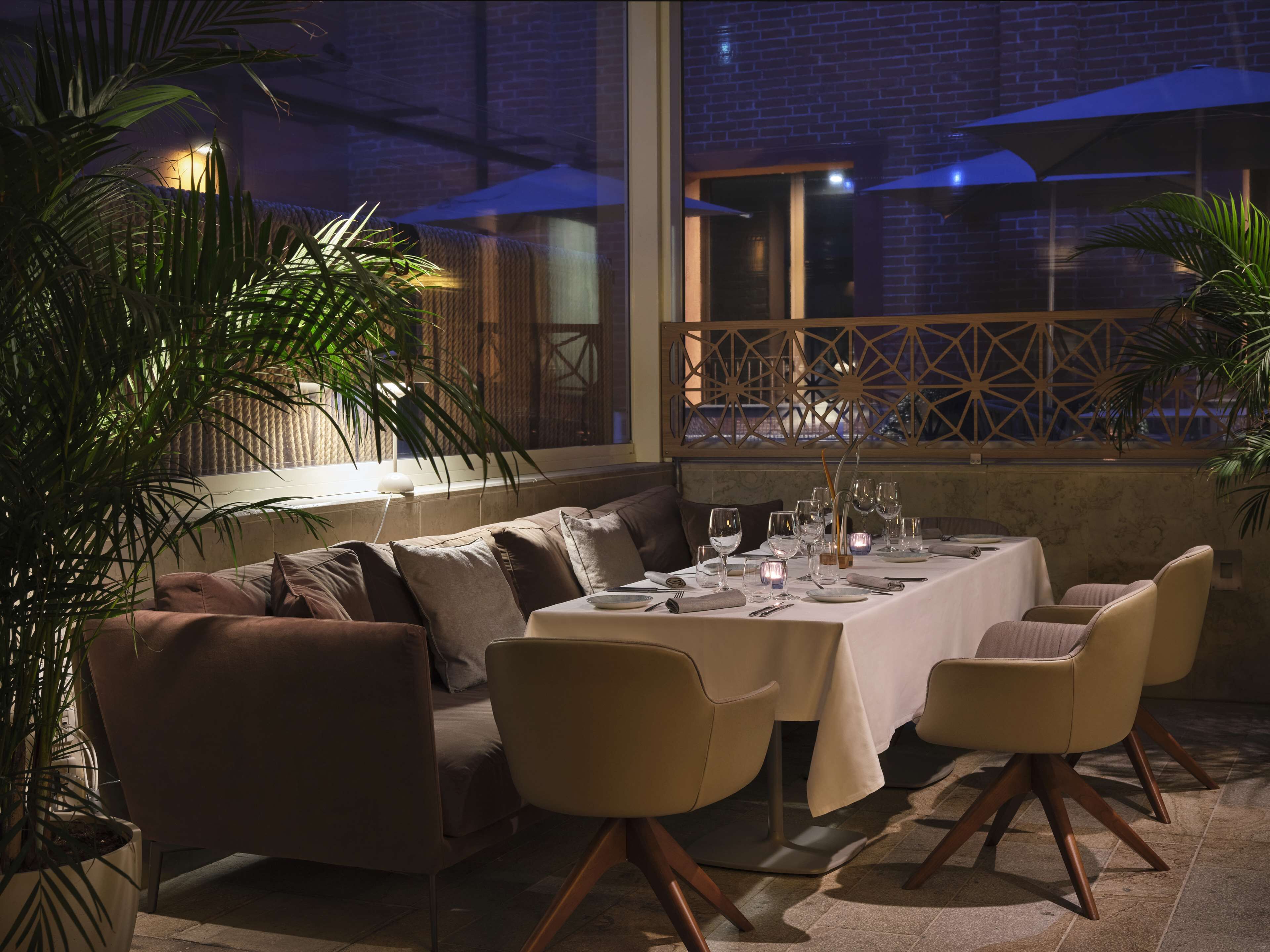 An outdoor dining area with a table set for four, surrounded by cushioned seating and potted plants.
