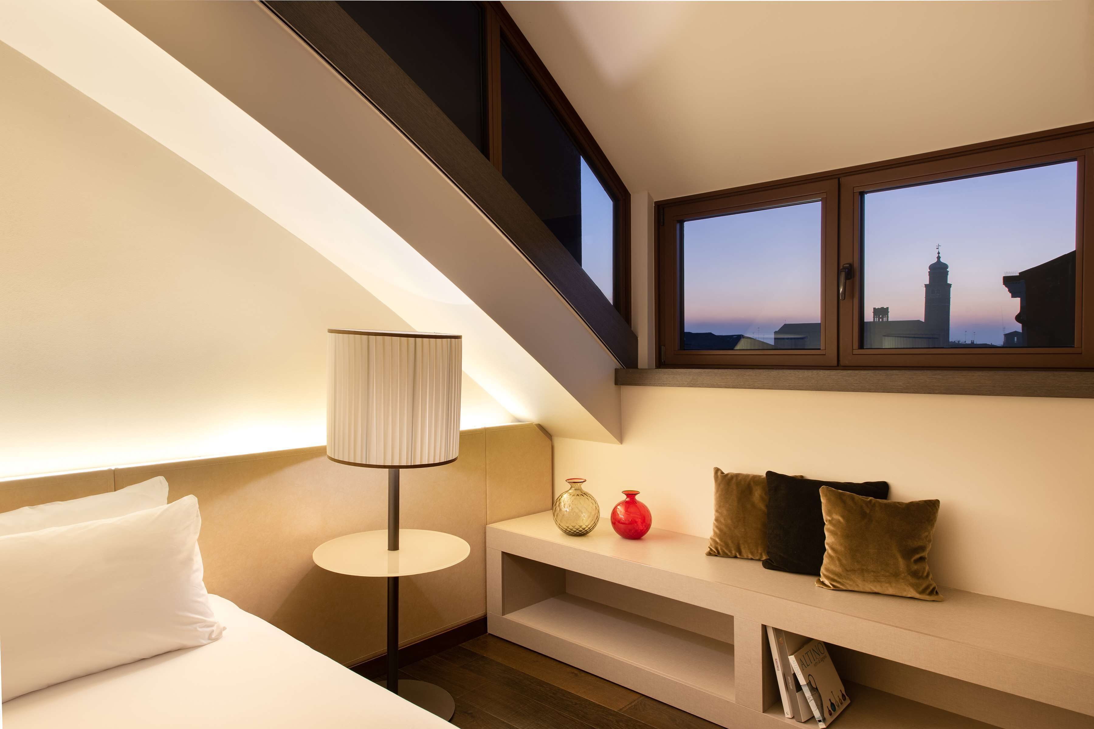 A hotel room with a bed, bedside lamp, shelf with decorative items, and a window with a cityscape view.