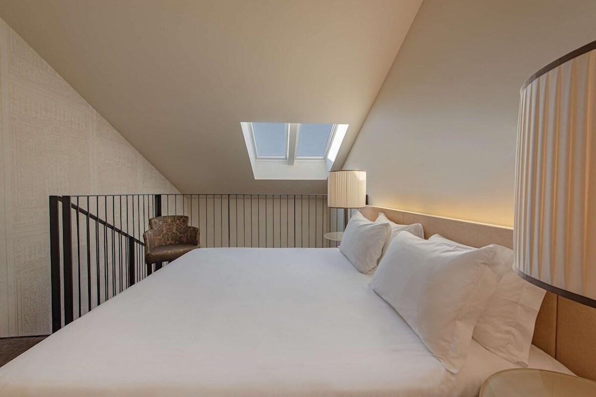 Un dormitorio con tragaluz, una cama con ropa de cama blanca, una mesita de noche con lámpara y una silla.