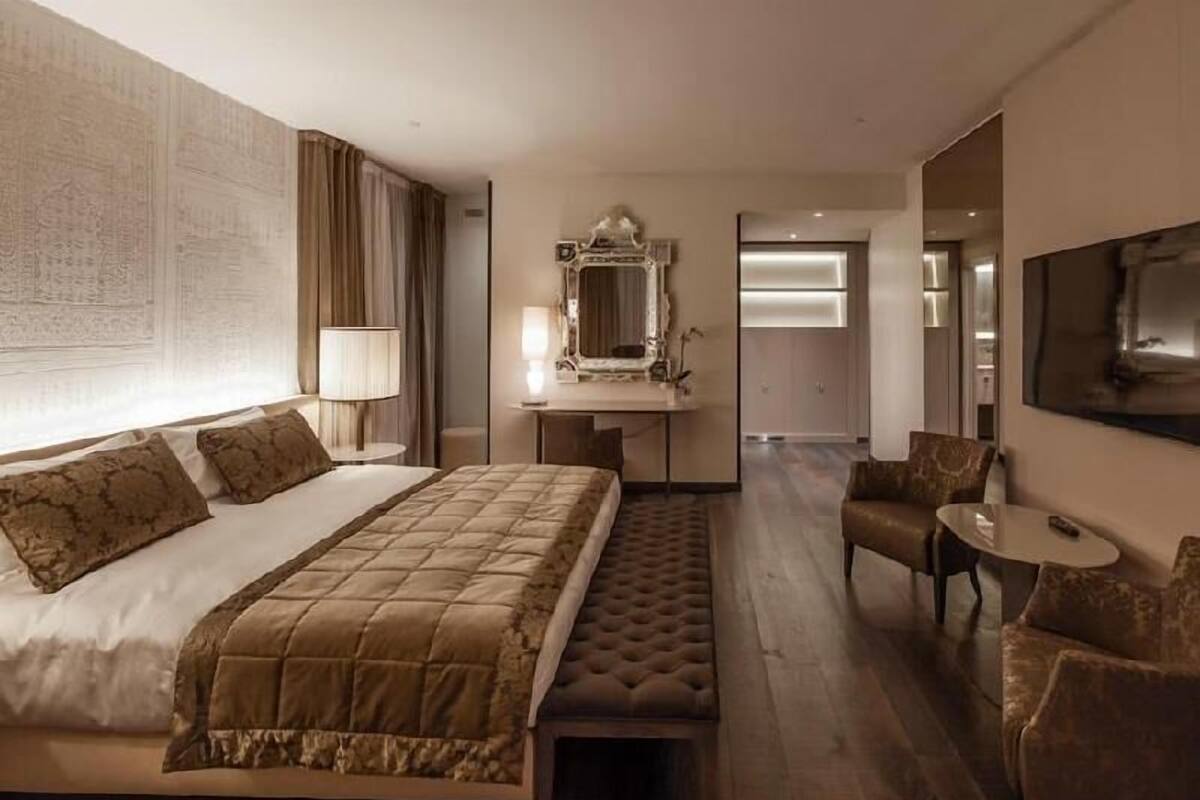 A hotel room with a large bed, a sitting area with a chair and a small table, a desk with a mirror, and a television mounted on the wall.